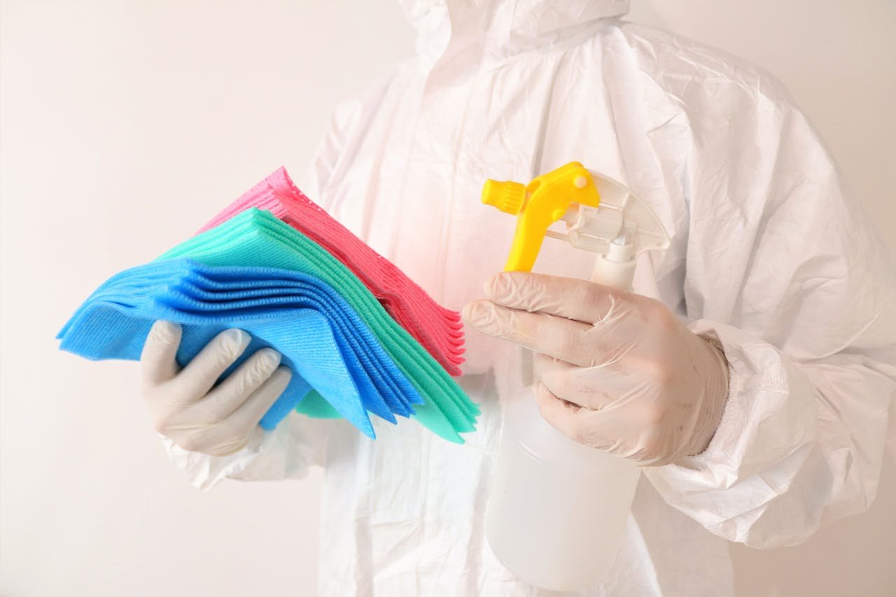 Individual in PPE holds cleaning cloths and spray bottle, ensuring hygiene and safety.